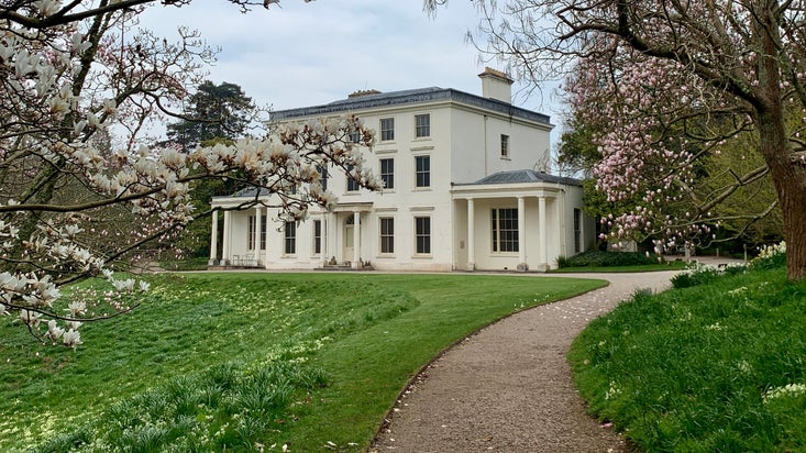 White manor house with gravel front and sloping green lawn, surrounded by magnolia trees with pink flowers.